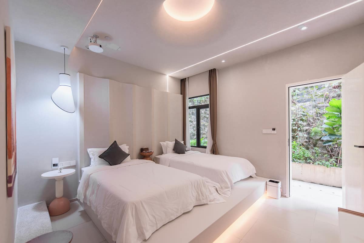 A modern bedroom with two beds, a bedside table, a small round table, and a window with a view of greenery.