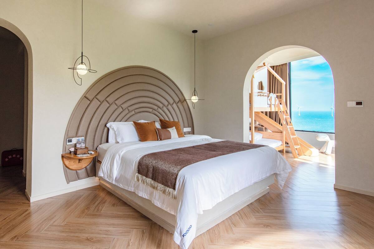 A spacious bedroom with a large bed, wooden headboard, and a view of the sea through an arched doorway.