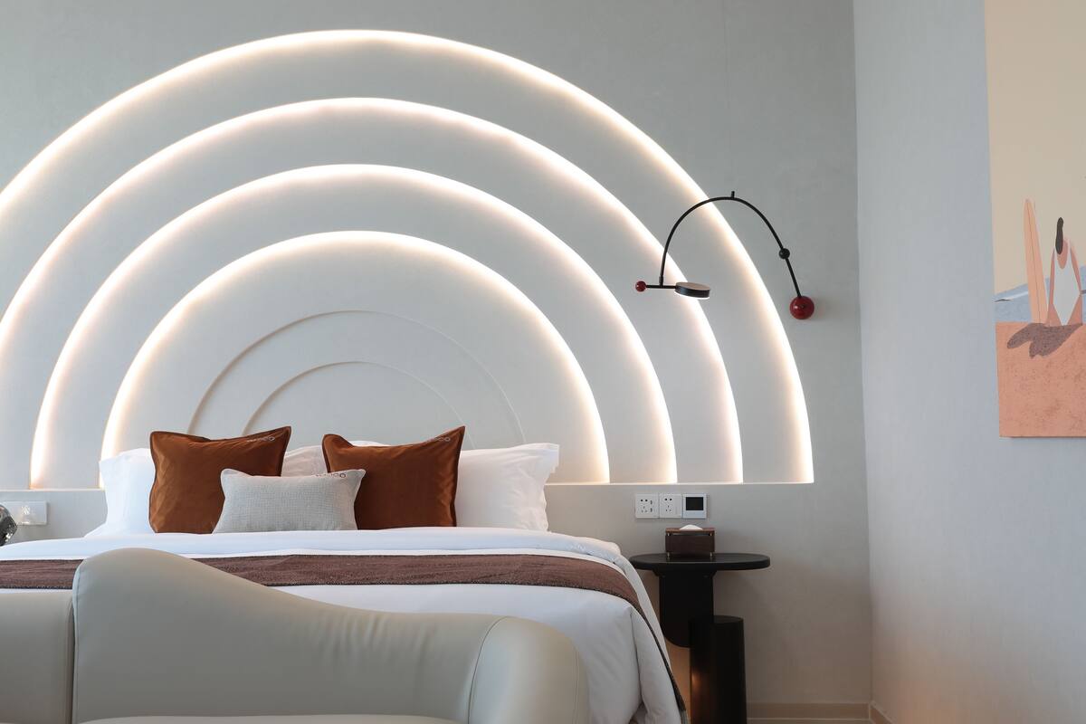 A modern hotel room with a curved ceiling light fixture, a bed with white and brown bedding, and a small bedside table with a lamp.