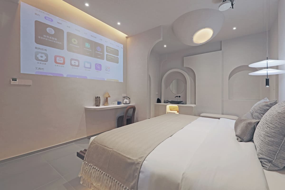 A modern hotel room with a large bed, a bedside table, a mirror, and a projection screen displaying room information.