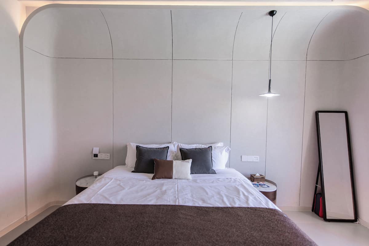 A modern hotel room with a large bed, a bedside table, a nightstand, a mirror, and a hanging light fixture.