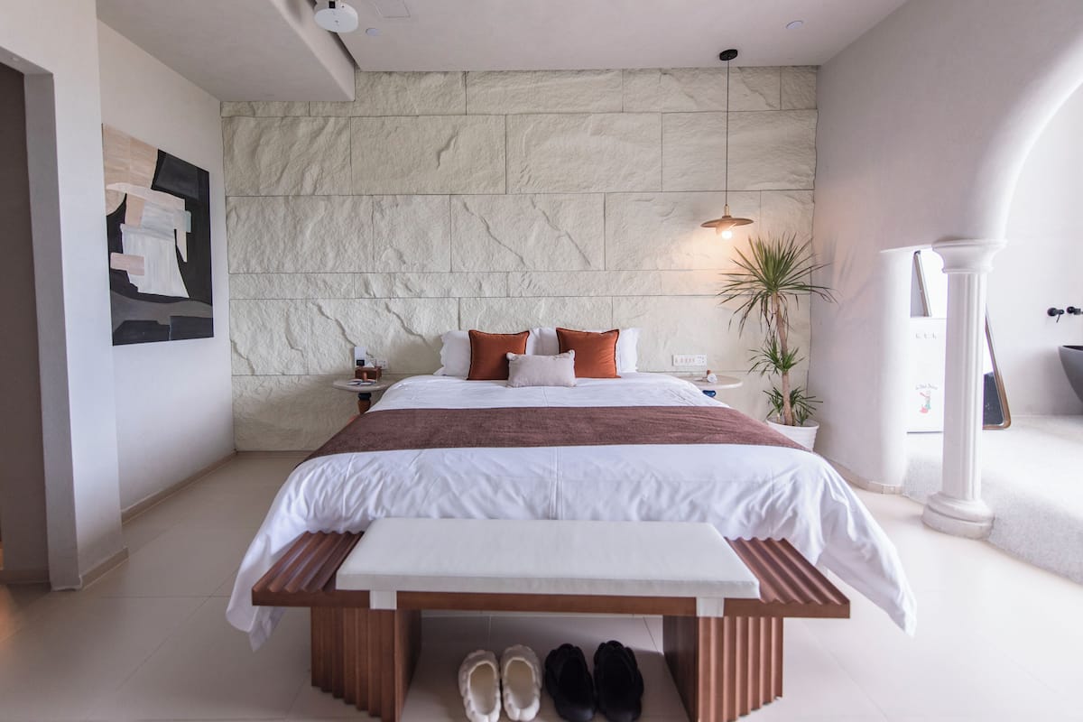 A modern bedroom with a large bed, a bench, a nightstand, and a potted plant.