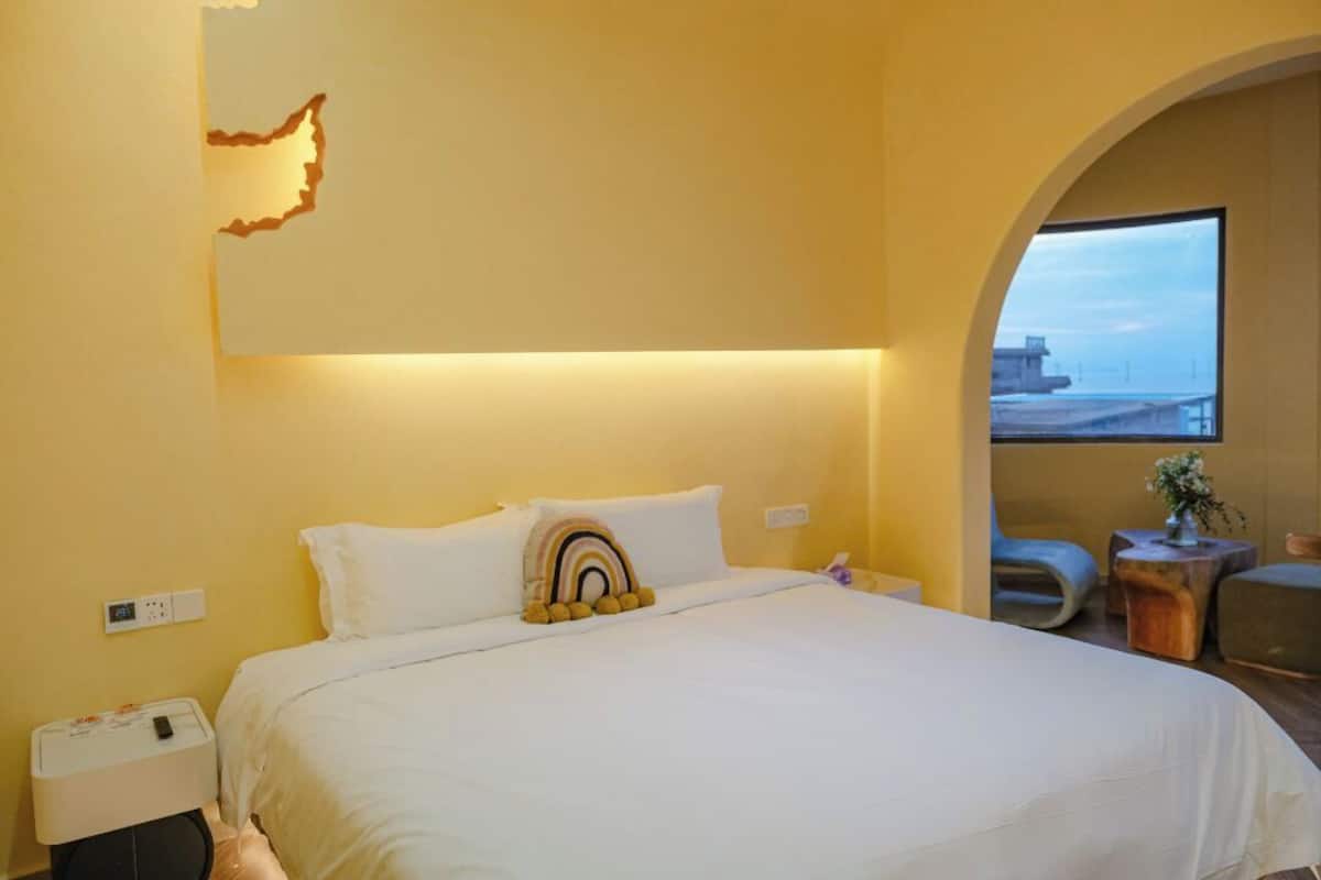 A hotel room with a large bed, a nightstand, a small table with a plant, and a view of the sea through an arched window.