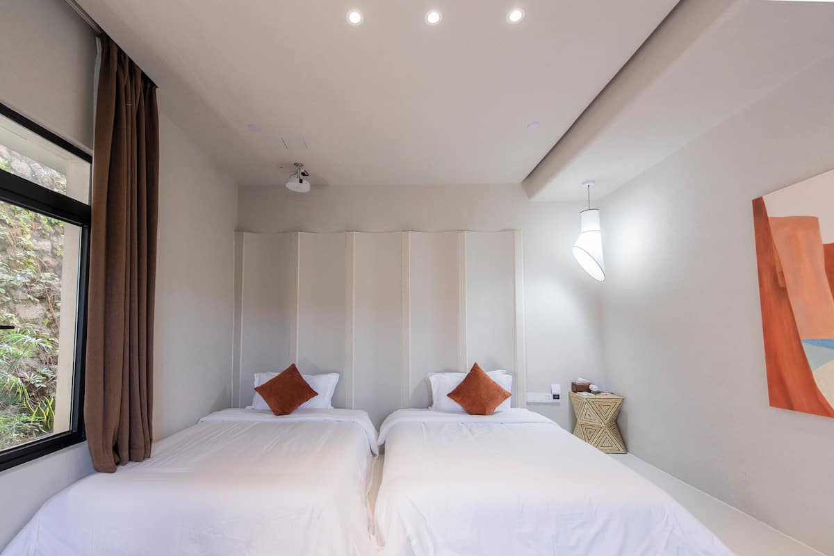 A hotel room with two beds, a window with curtains, a ceiling fan, a wall-mounted light, and a small side table.