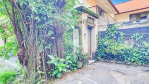 A courtyard with dense foliage, a concrete pathway, and a building with a tiled roof and a balcony. - Collection O Putri Inn Homestay (Denpasar)