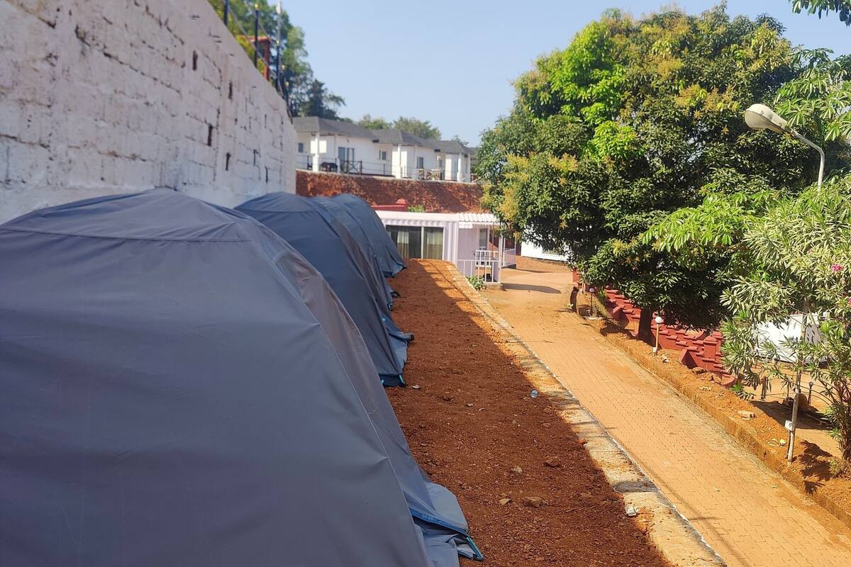 A row of tents along a dirt path with a white brick wall on one side and a tree-lined area on the other.