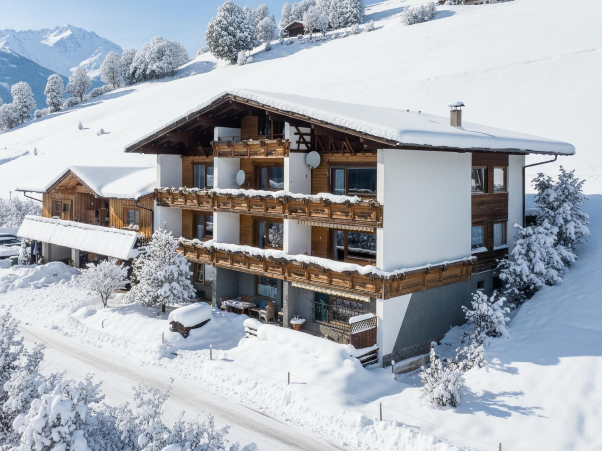 A multi-story chalet with a wooden facade and balconies, surrounded by snow-covered trees and a mountain backdrop.