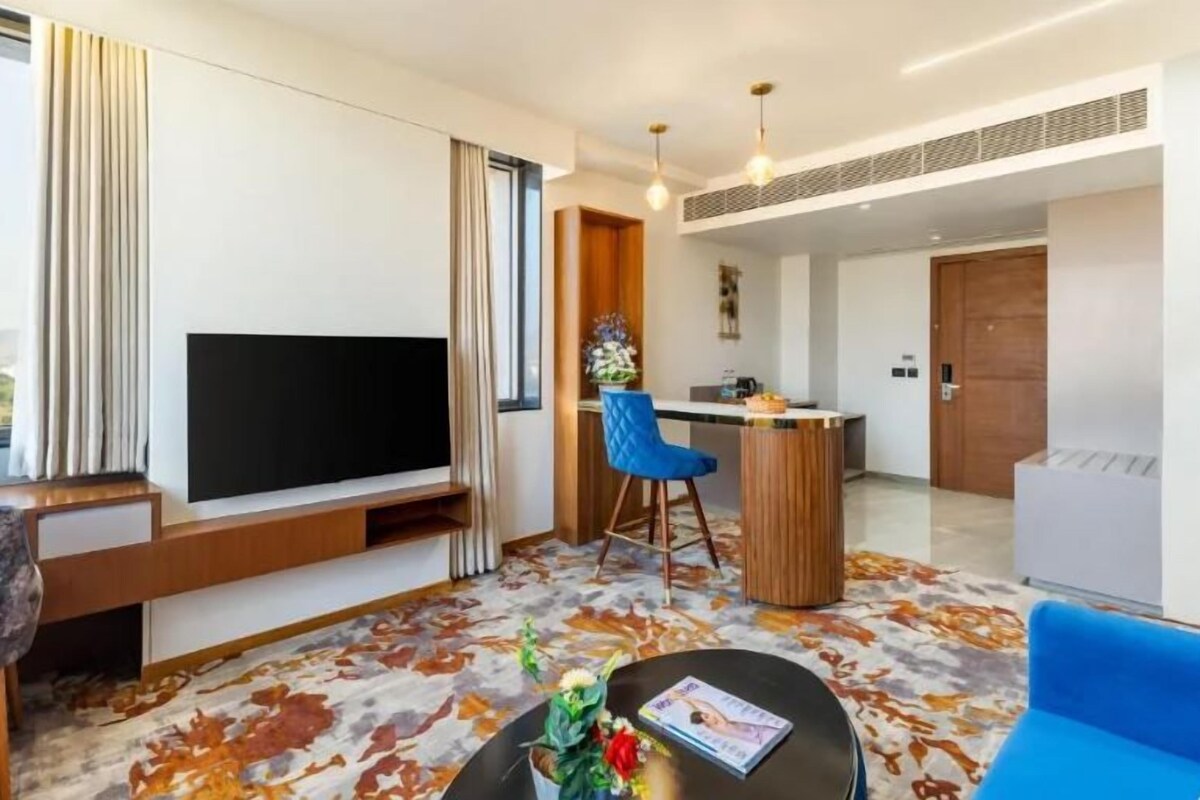 A modern hotel room with a large flat-screen TV, a dining area with a bar, and a view of the outdoors.