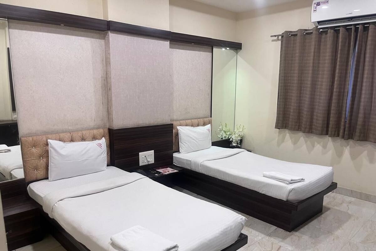 A hotel room with two single beds, a large mirror, a window with curtains, and a wall-mounted air conditioner.
