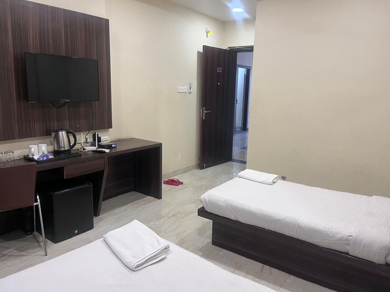 A hotel room with two beds, a desk with a computer, a flat-screen TV mounted on the wall, and a small kitchenette area.