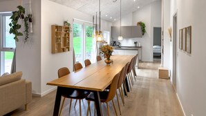 A modern dining area with a wooden table and chairs, a kitchen in the background, and a view of the outdoors through a large window. - Lovely home in Ebeltoft (Ebeltoft)