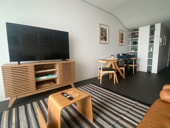 A modern living room with a flat-screen TV, wooden entertainment unit, dining table with chairs, and a striped rug.