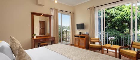 Habitación de hotel con cama, escritorio, silla, televisor y balcón con vistas a zonas verdes.