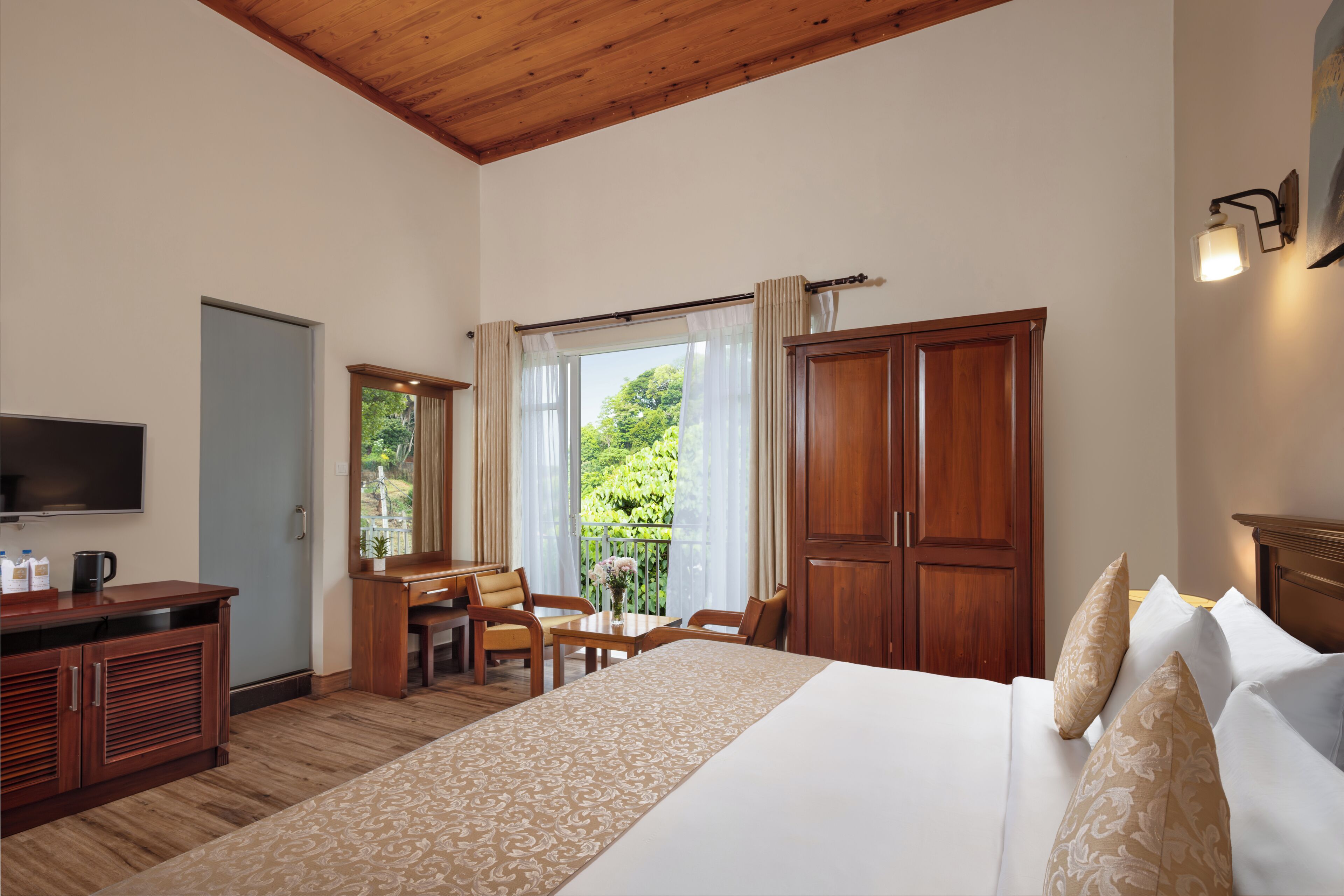 A hotel room with a bed, a desk, a television, and a view of greenery outside.