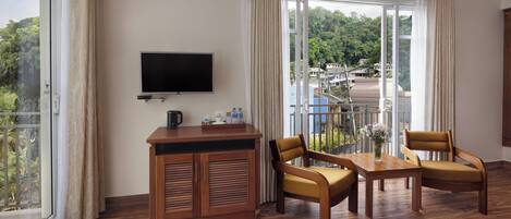 Habitación de hotel con televisión de pantalla plana, muebles de madera, una mesa con un florero y un balcón con vistas al follaje.