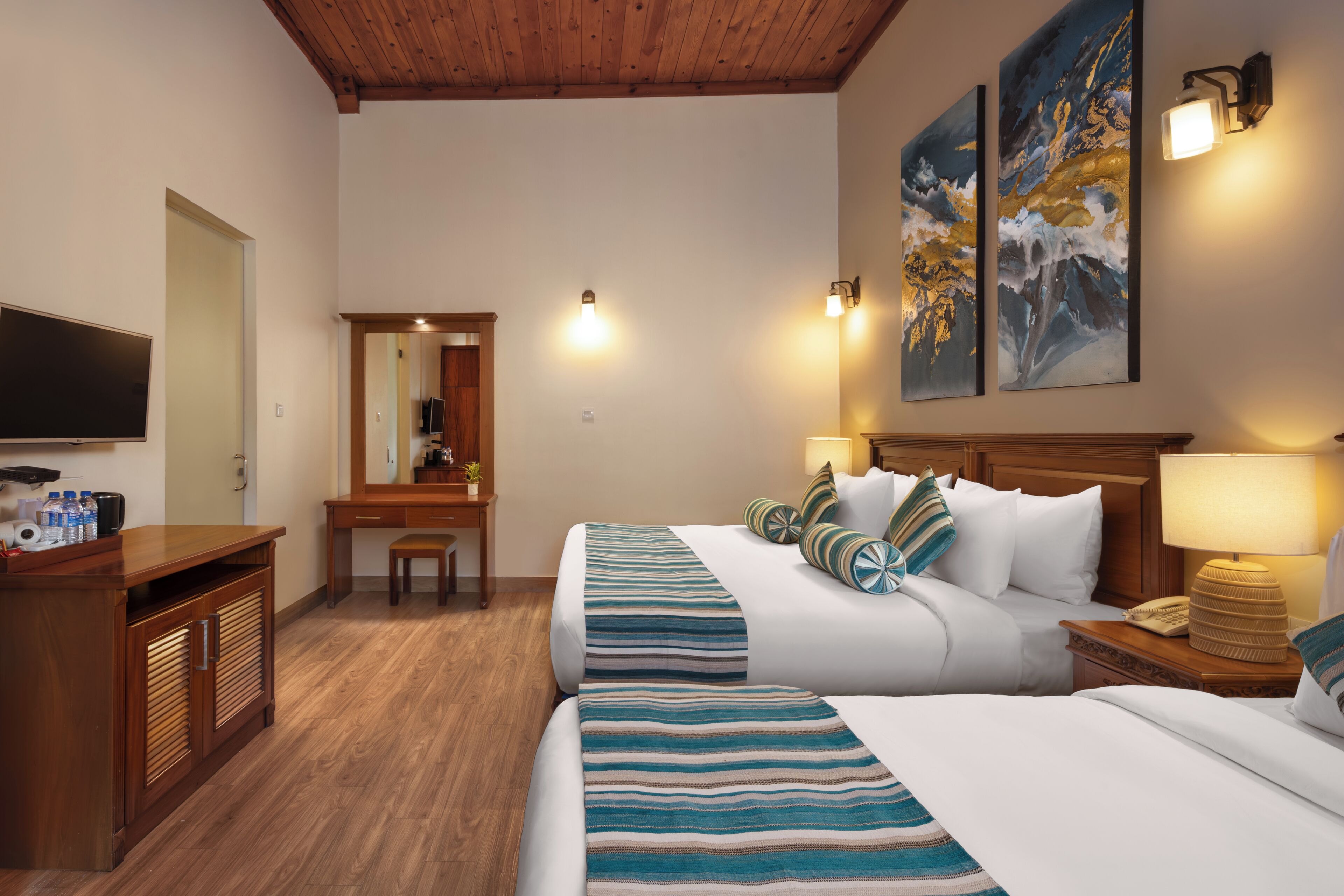 A hotel room with two beds, a desk, a television, and artwork on the walls.