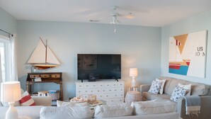A living room with a TV, white furniture, a ceiling fan, and a sailboat painting.