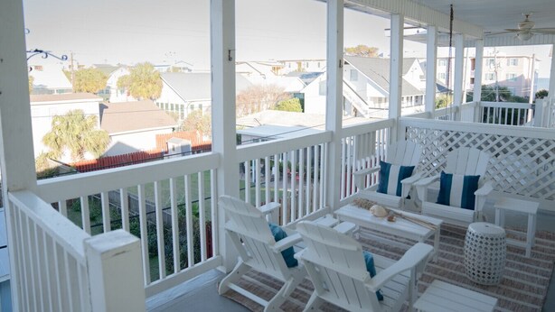 A covered outdoor seating area with white chairs and a table, overlooking a residential neighborhood.