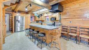 A wooden cabin interior with a kitchen featuring a stainless steel refrigerator, a microwave, and a large island with bar stools. - < 1 Mi to Kenai River: Spacious Cabin w/ Deck! (Sterling)