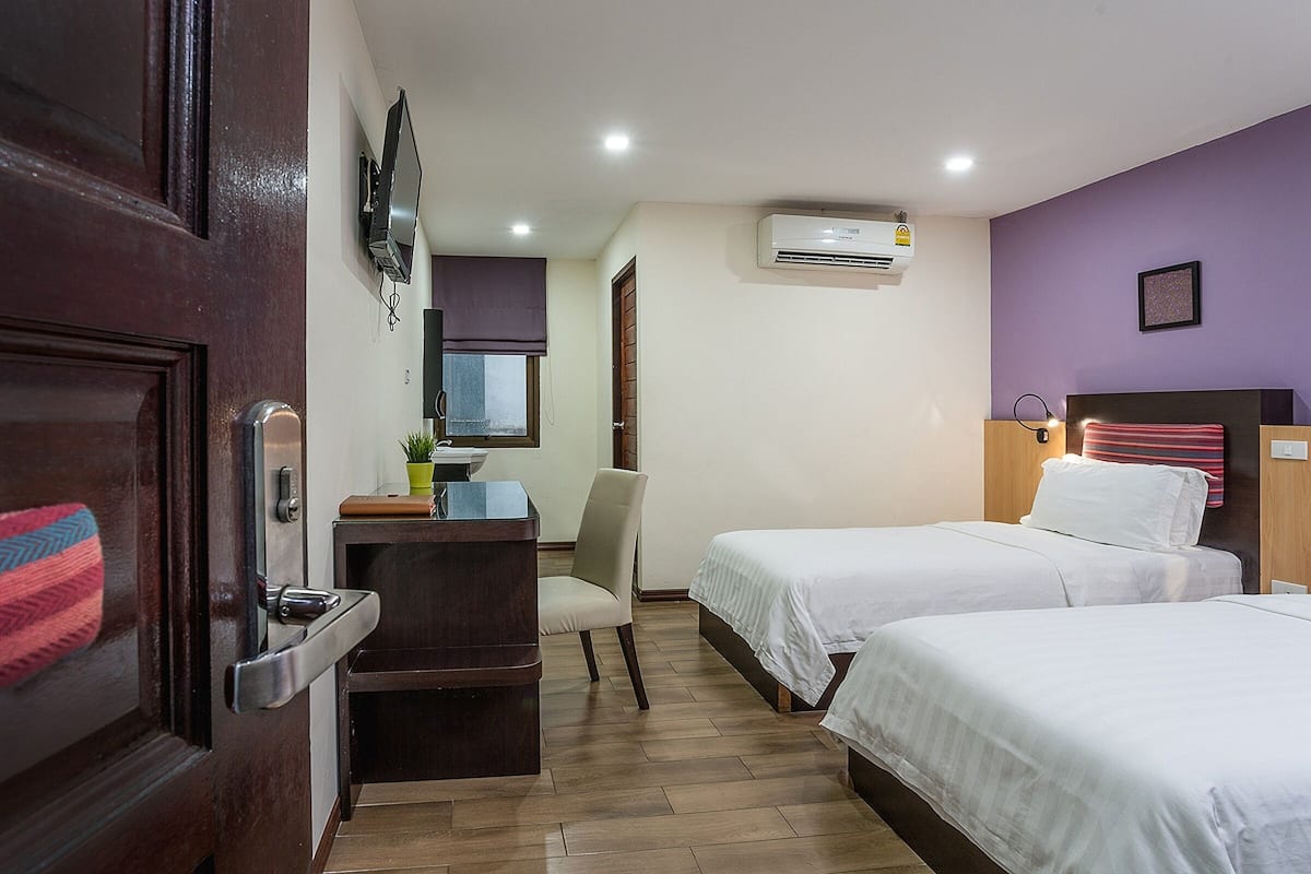 A hotel room with two beds, a desk, a chair, a TV, and an air conditioning unit.