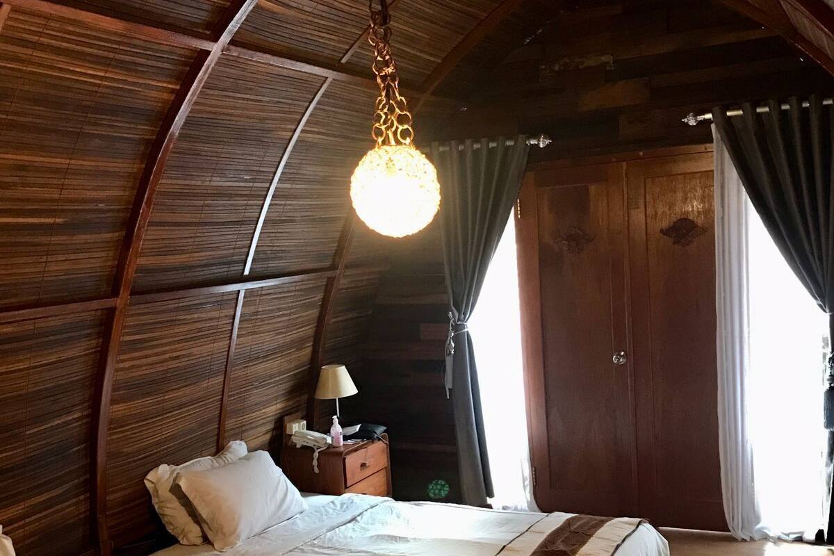 Une chambre à coucher chaleureuse et rustique avec un plafond voûté, deux lits avec une literie à rayures, un luminaire suspendu et une table de chevet avec une lampe.