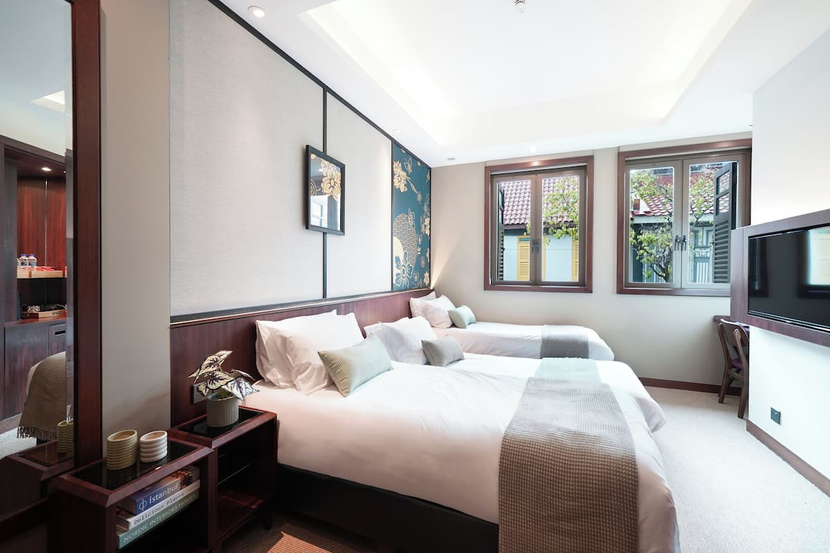 Quarto de hotel com duas camas, televisão, secretária com candeeiro e janela com vista para áreas verdes.