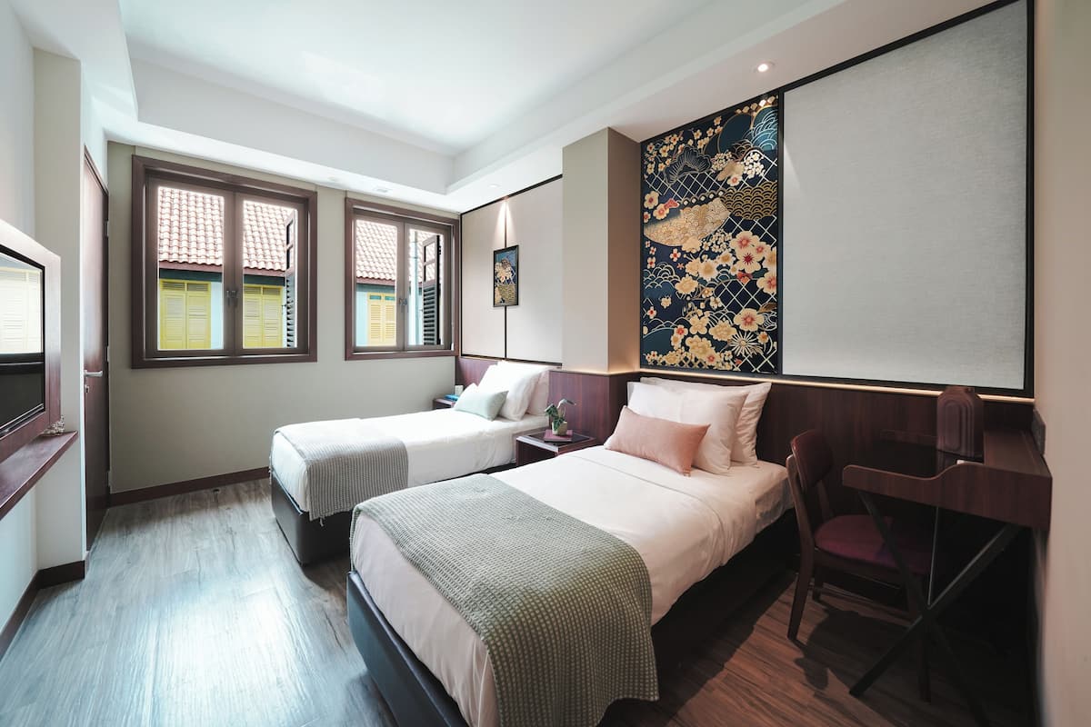 Quarto de hotel com duas camas, uma escrivaninha e uma cadeira. Há janelas com persianas, uma parede com painel decorativo e uma televisão à esquerda.