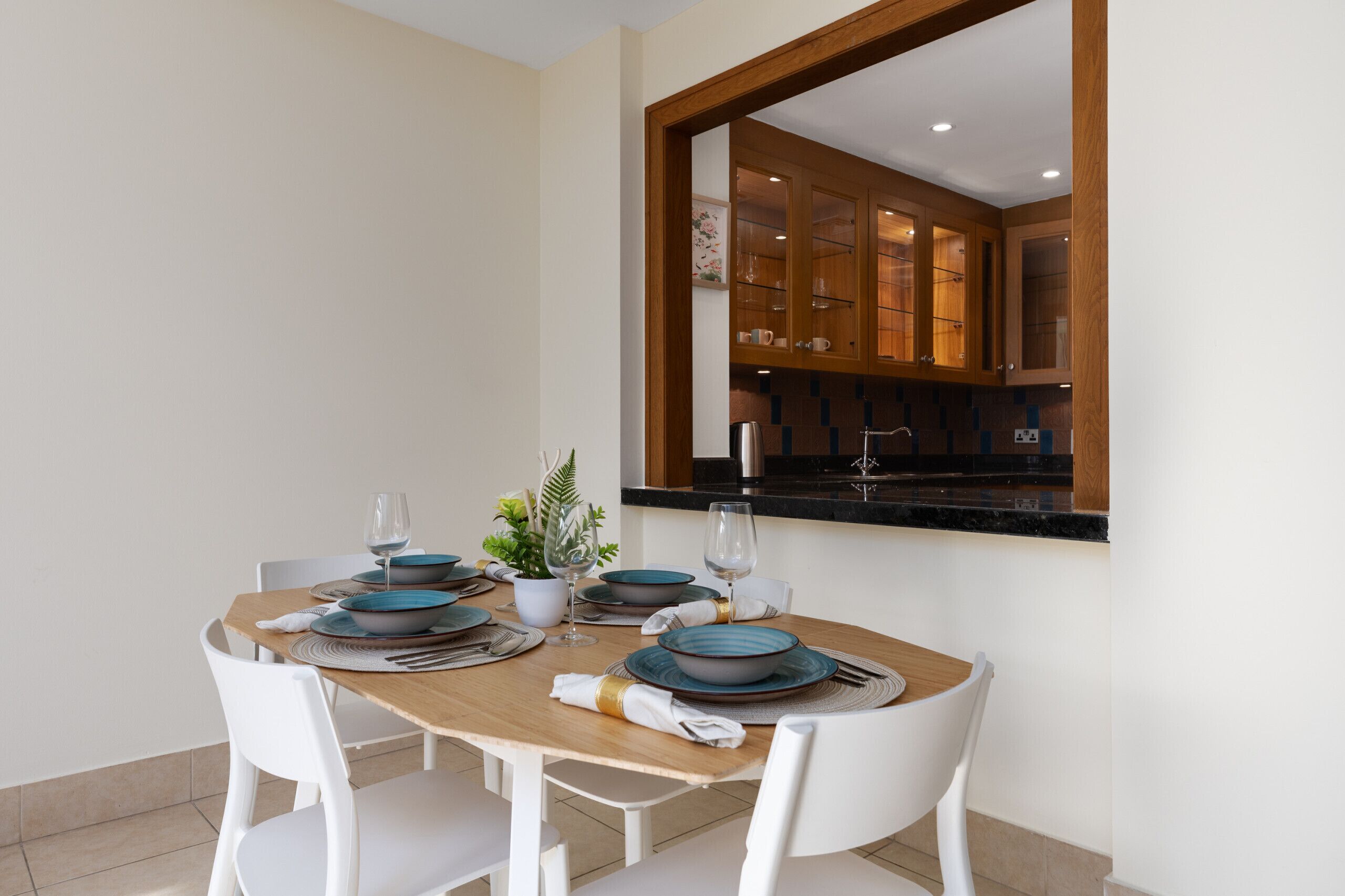 Un coin repas avec une table en bois dressée pour quatre personnes, des chaises blanches et une vue sur la cuisine à travers une fenêtre à cadre en bois.