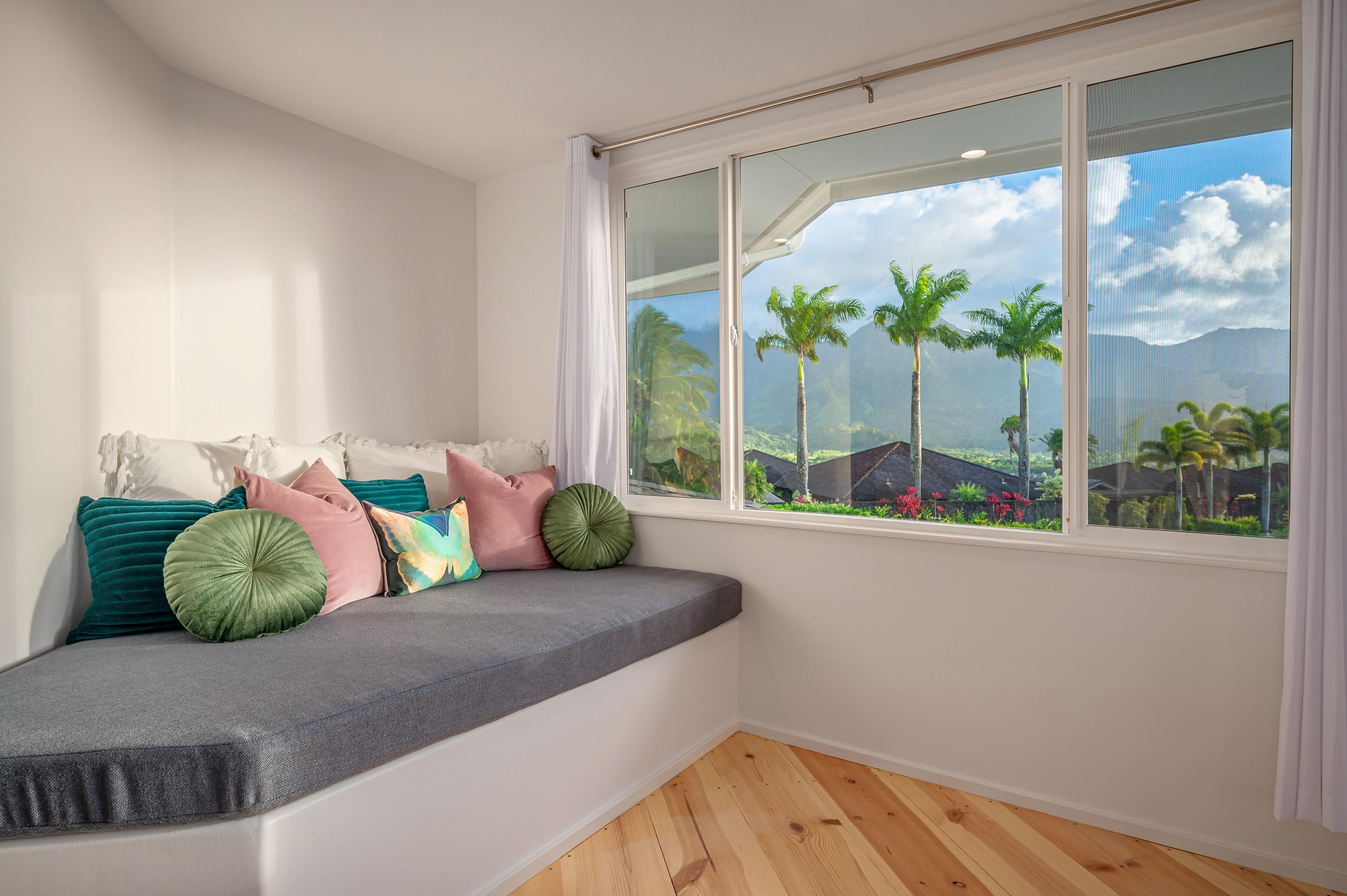 A room with a built-in bench against a wall, a large window with a view of palm trees and mountains, and a wooden floor.