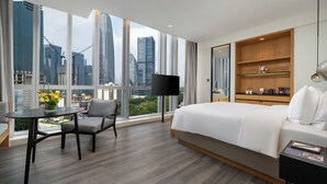 Un cuarto de hotel con una cama grande, un televisor, un estante de madera, una mesita con una silla y vistas a edificios de la ciudad.