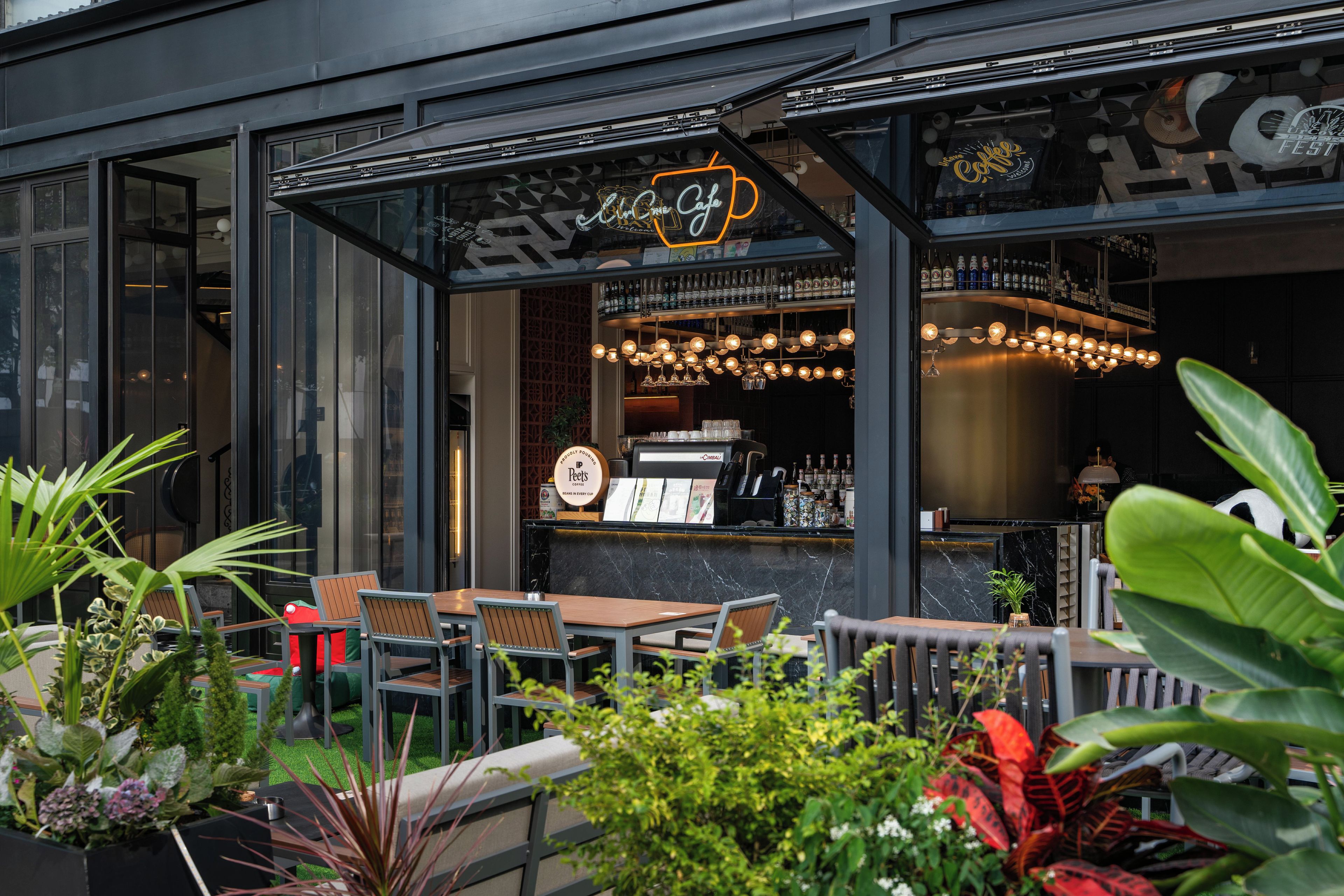 An outdoor seating area with tables and chairs, a coffee shop with a visible counter and shelves of bottles, and a large illuminated sign above the entrance.
