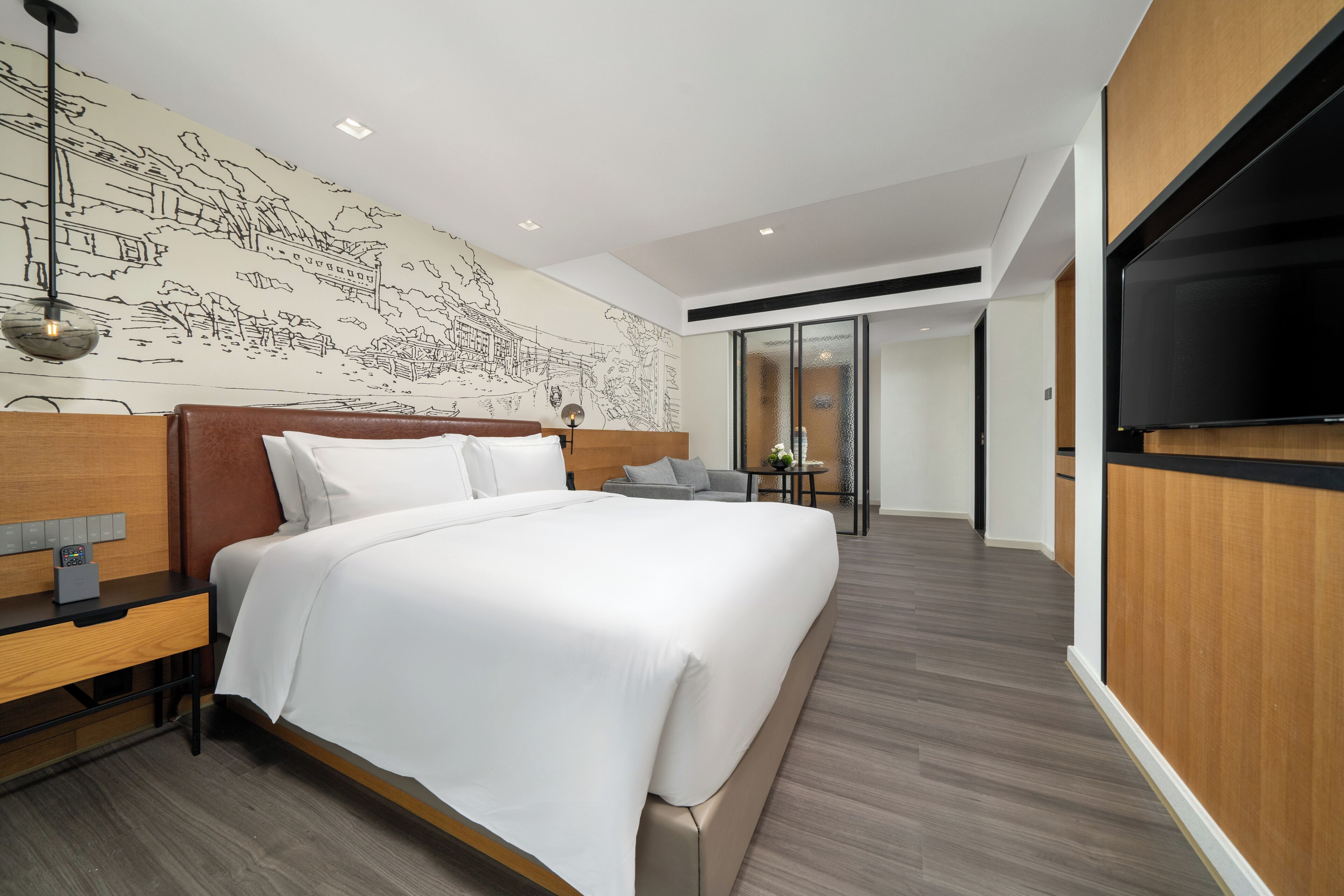 A modern hotel room with a large bed, a wooden headboard, a desk with a lamp, a wall mural, and a TV.