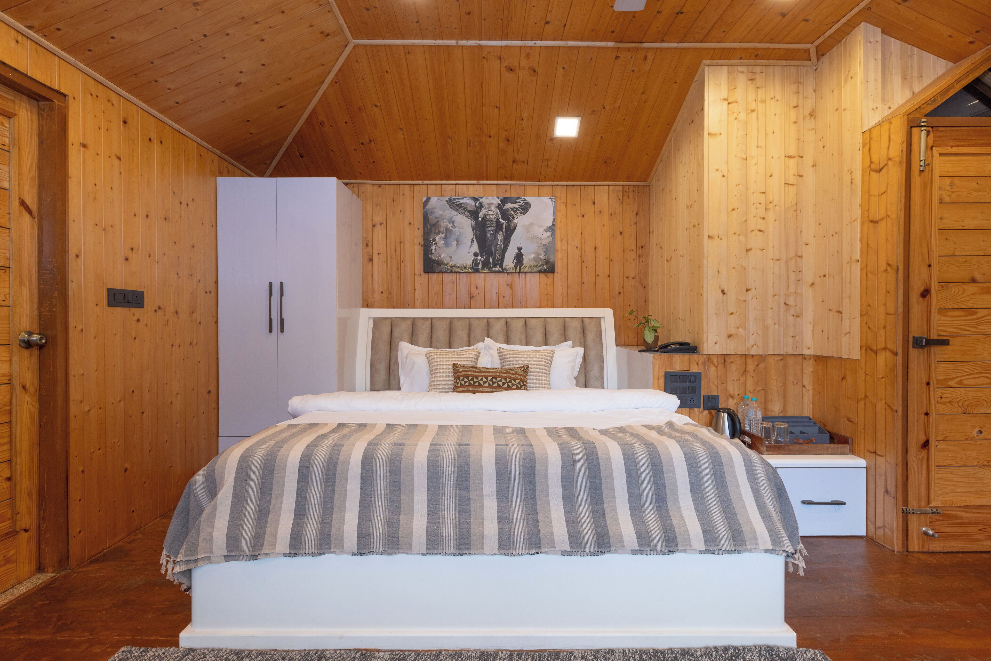A bedroom with a wooden ceiling, a bed with a striped blanket, a nightstand with a lamp, and a painting on the wall.