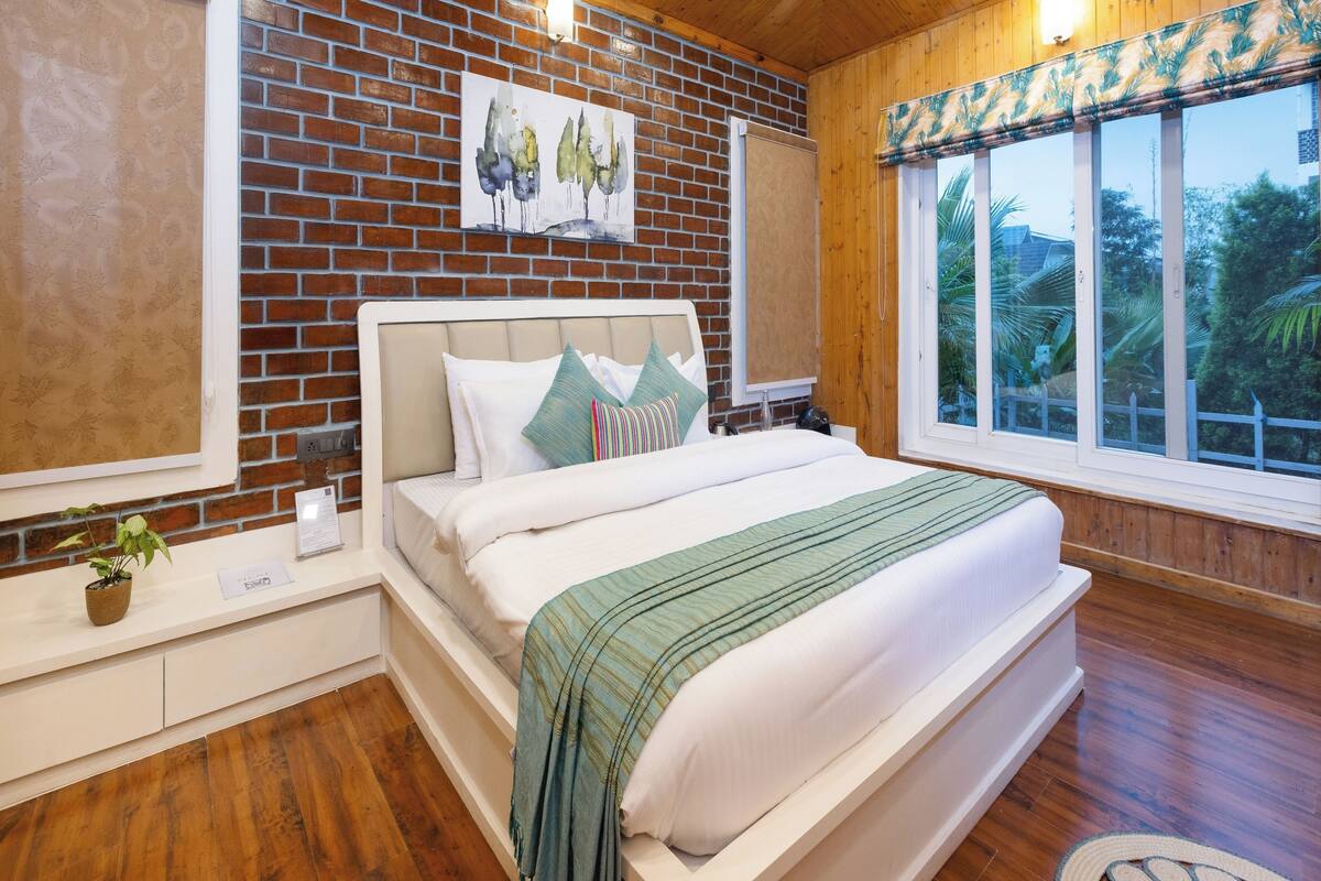 A bedroom with a brick wall, a wooden ceiling, a bed with white bedding and a green blanket, a nightstand, and a window with a view of trees.