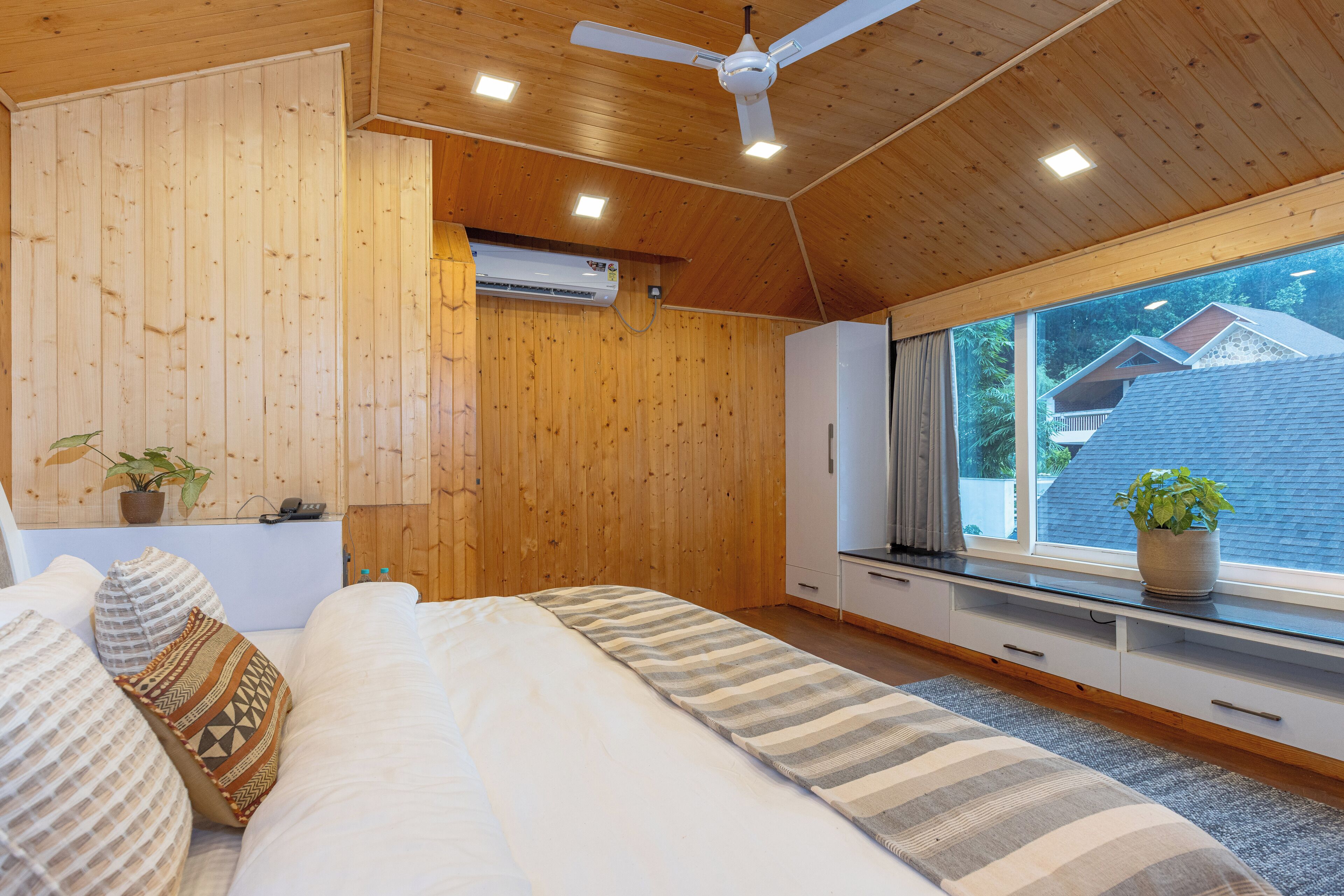 A bedroom with a bed, a ceiling fan, an air conditioning unit, a window with a view of houses, and a plant on the windowsill.