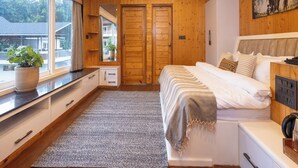A bedroom with wooden walls and ceiling, a bed with white bedding, a ceiling fan, a large window with a view of a house, and a potted plant on the windowsill. - Megha Resort & Villas, Dehradun-Where Nature Pampers You (Vikasnagar)