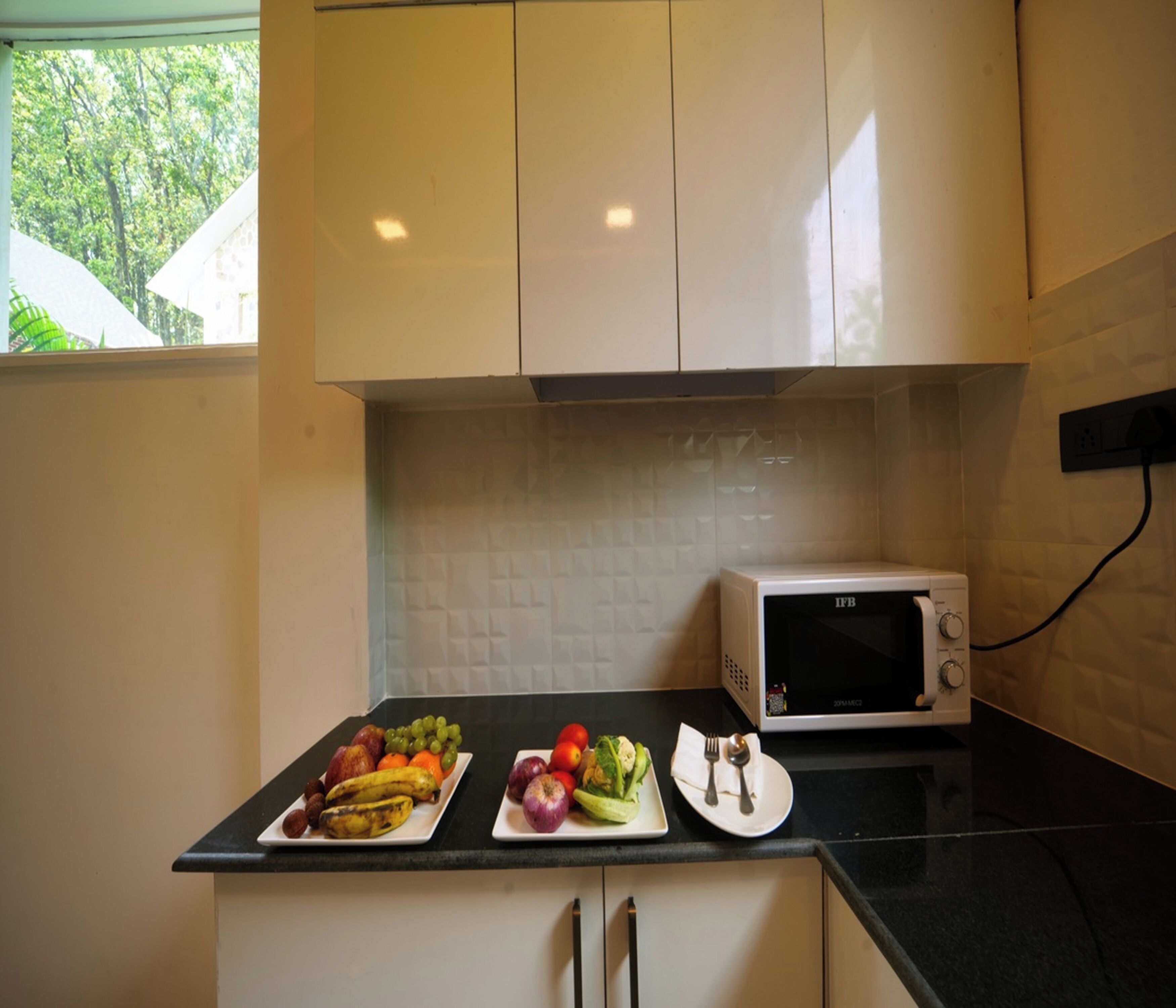 Een keuken met witte kasten, een zwart aanrecht en een witte magnetron. Op het aanrecht staan schalen met fruit en groenten.