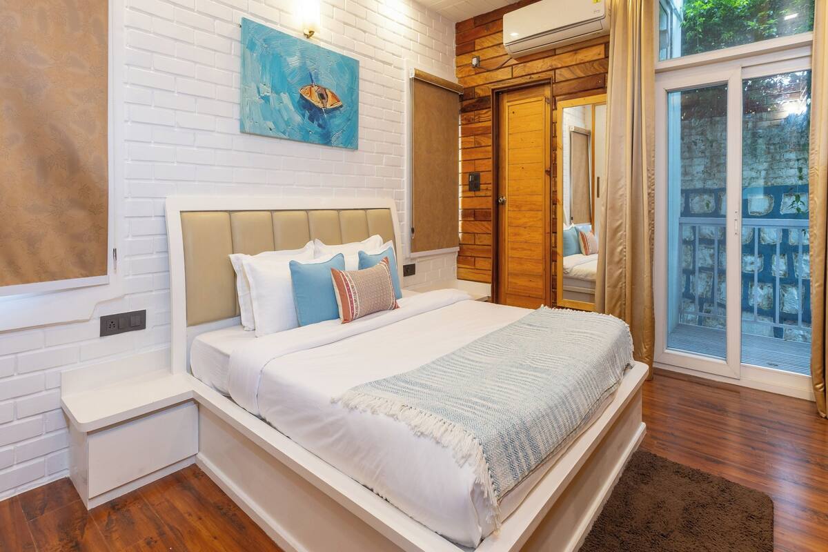 A modern bedroom with a large bed, a wooden wardrobe, a white brick wall, and a large window with a view of greenery.