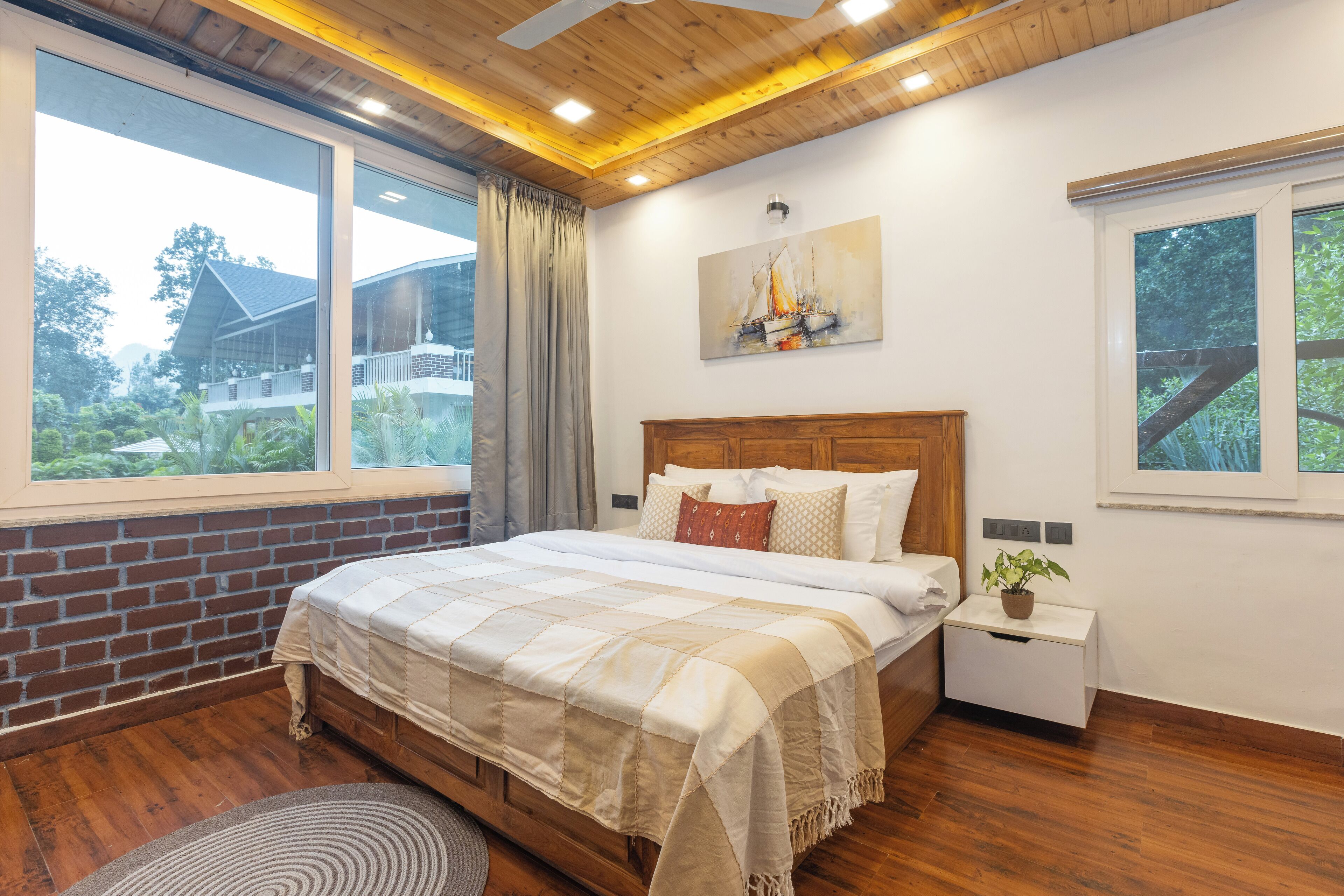 A bedroom with a large bed, a nightstand with a plant, and a window with a view of a house and greenery.