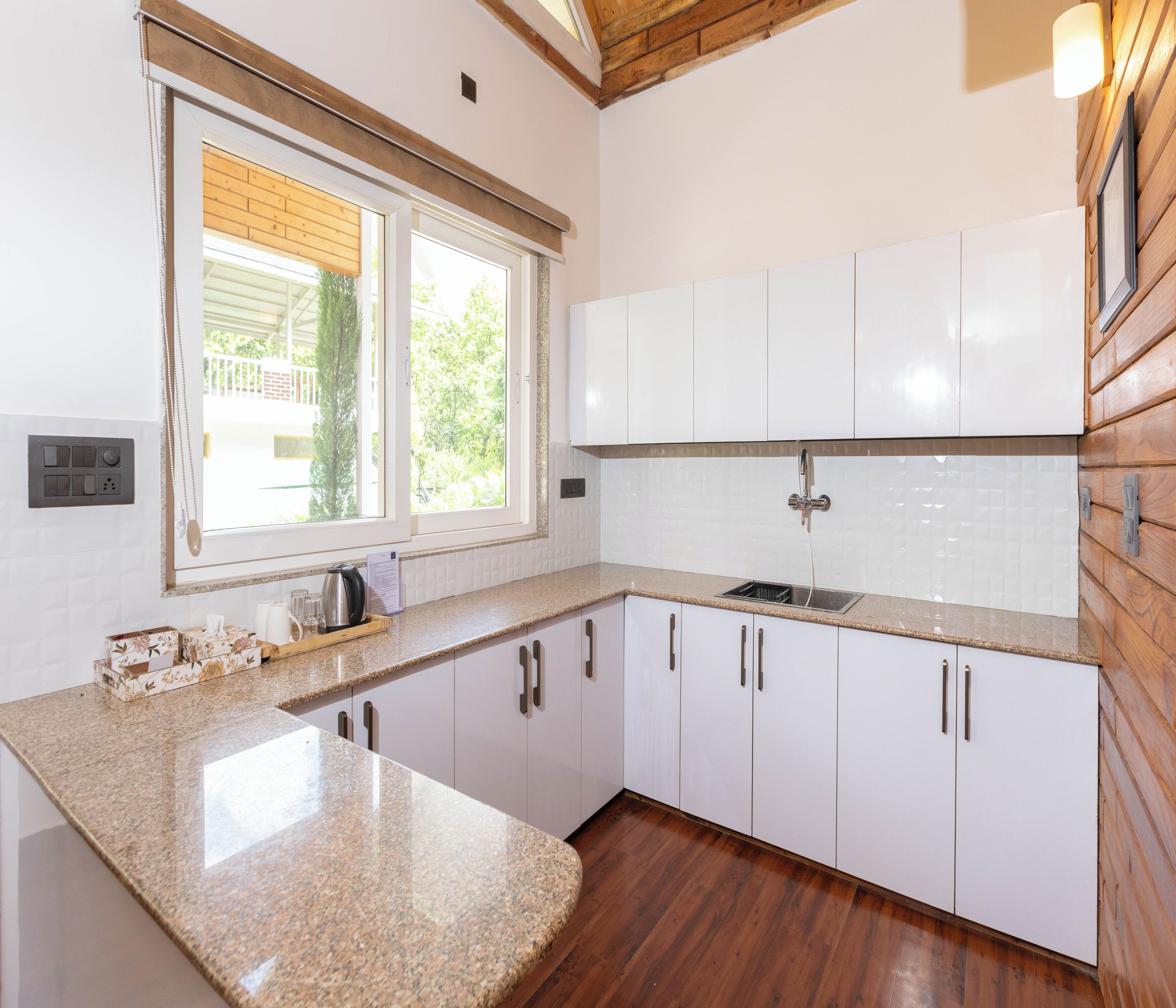 A kitchen with white cabinets, a granite countertop, a window with a view of greenery, and a wooden ceiling.