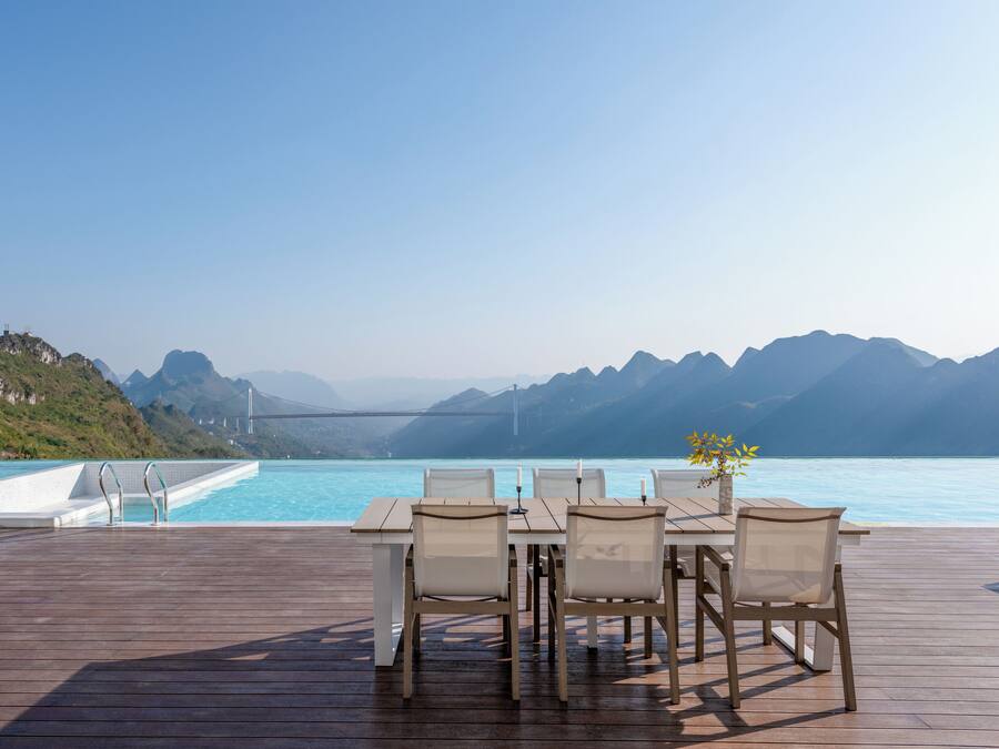 En träveranda med ett bord och stolar, med utsikt över en pool och berg.
