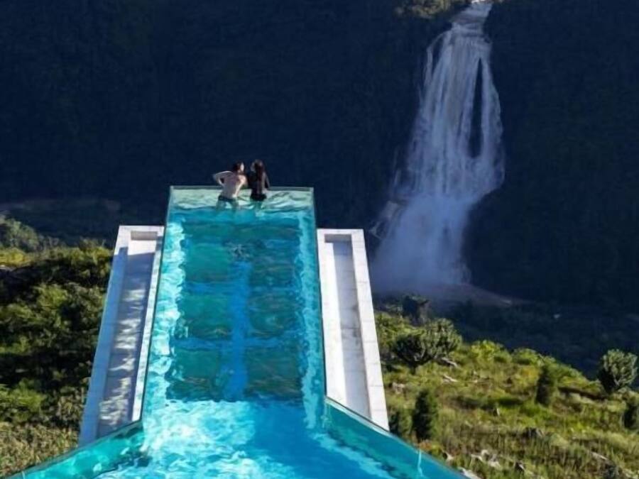 En rektangulär infinitypool med två personer i den bortre änden, med utsikt över ett vattenfall och frodig grönska.