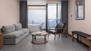 A modern hotel room with a grey sofa, a round wooden coffee table, a black leather chair, a wooden bench, and a large window with a view of the sea. - YAOJIA WANGPU VILLA (Anshun)