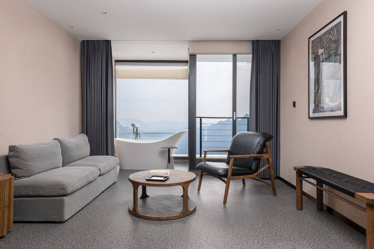 A modern hotel room with a grey sofa, a round wooden coffee table, a black leather chair, a wooden bench, and a large window with a view of the sea.