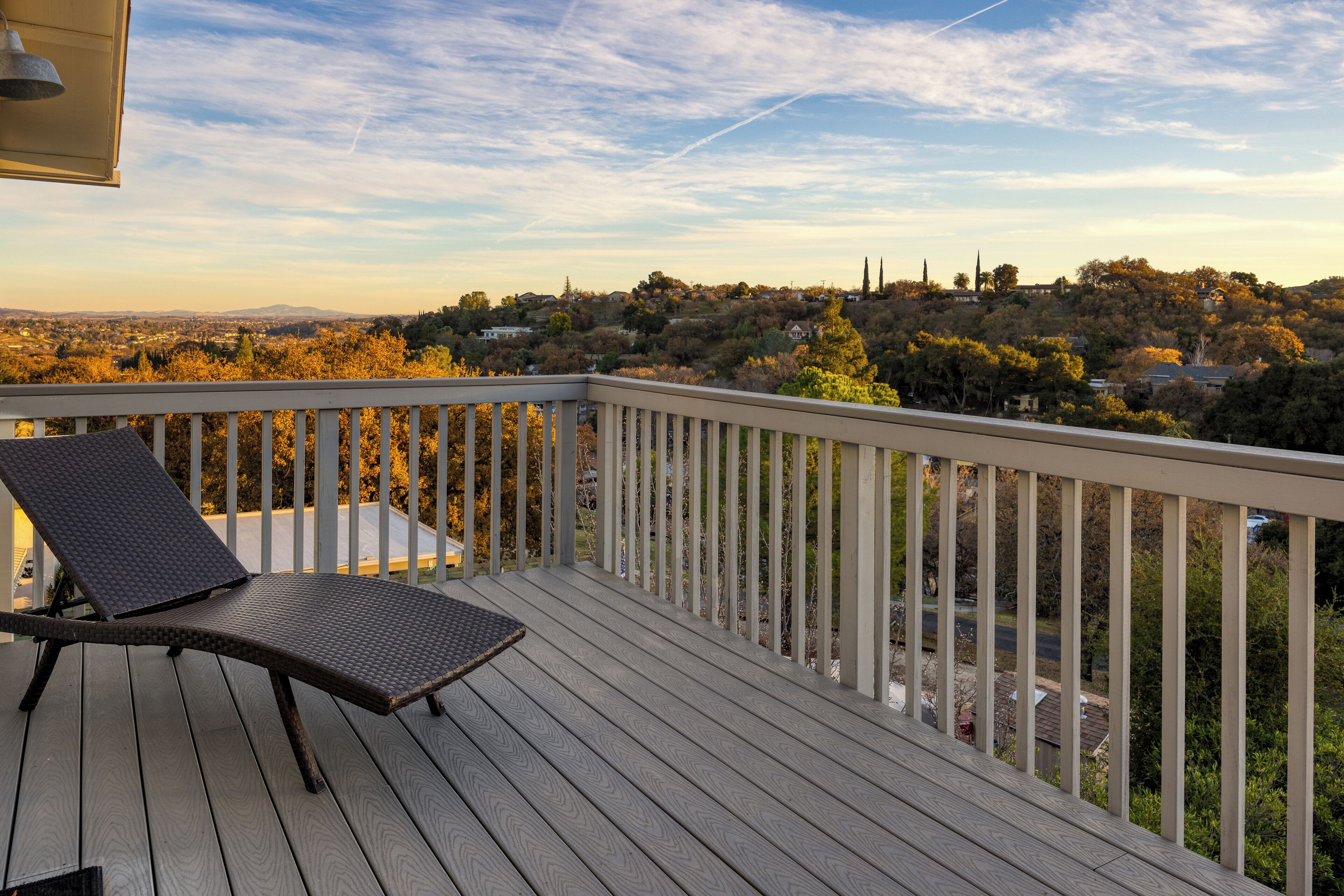 A wooden deck with a metal railing, a single lounge chair, and a view of a residential area with trees and houses.
