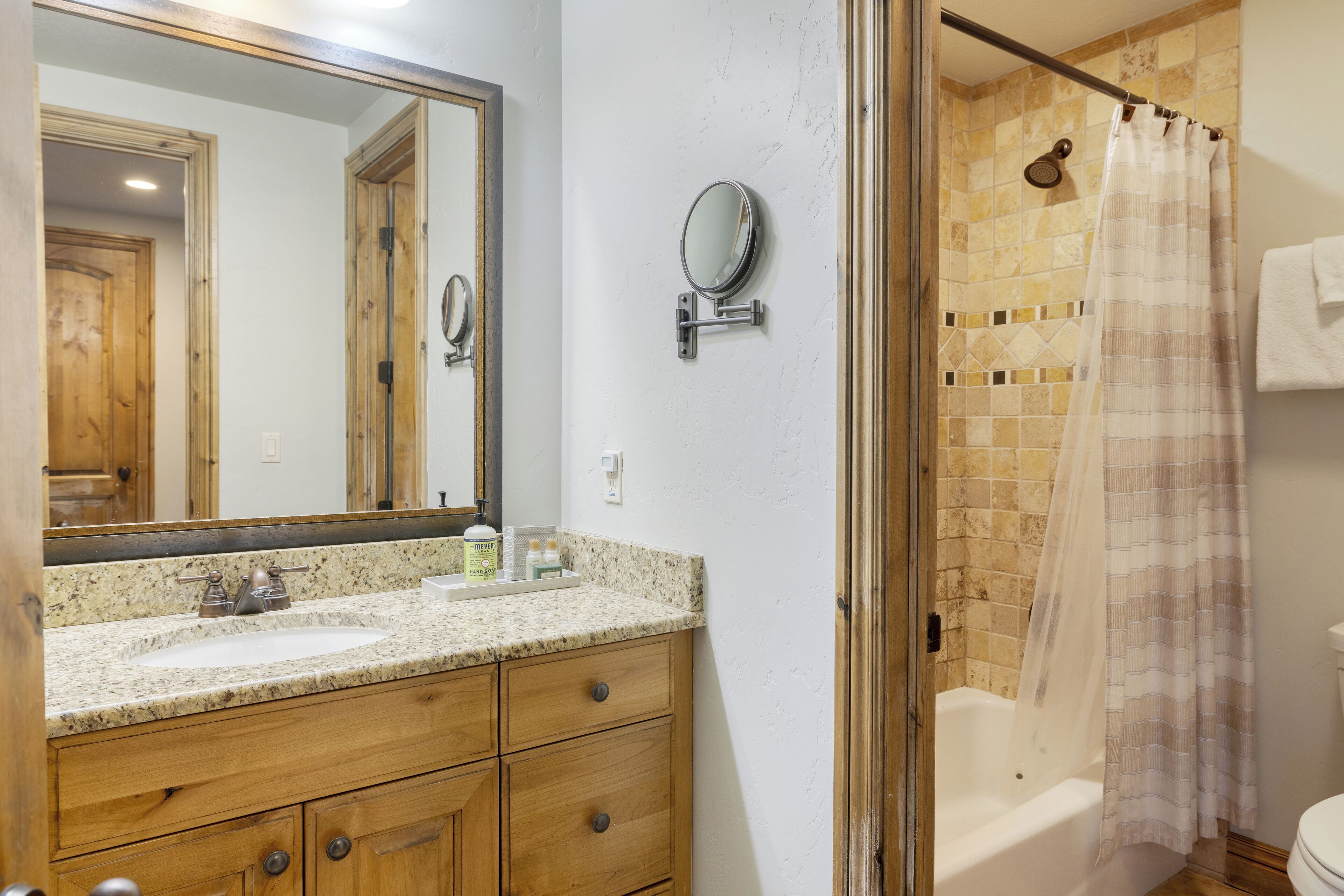 A bathroom with a granite countertop, wooden vanity, a large mirror, a small round mirror, a shower with a beige shower curtain, and a toilet.