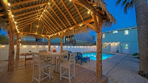 A wooden deck with a thatched roof and string lights, a pool, and a house in the background. - Seaglass Landing | Private Pool, Cabana, & Near Port Aransas Beaches (Port Aransas)