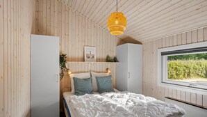 Ein Schlafzimmer mit Holzbalkendecke, einem Bett mit weißer Bettwäsche, einer Pendelleuchte, einem Fenster mit Blick ins Grüne und einem Regal mit Pflanzen und einem Bild.