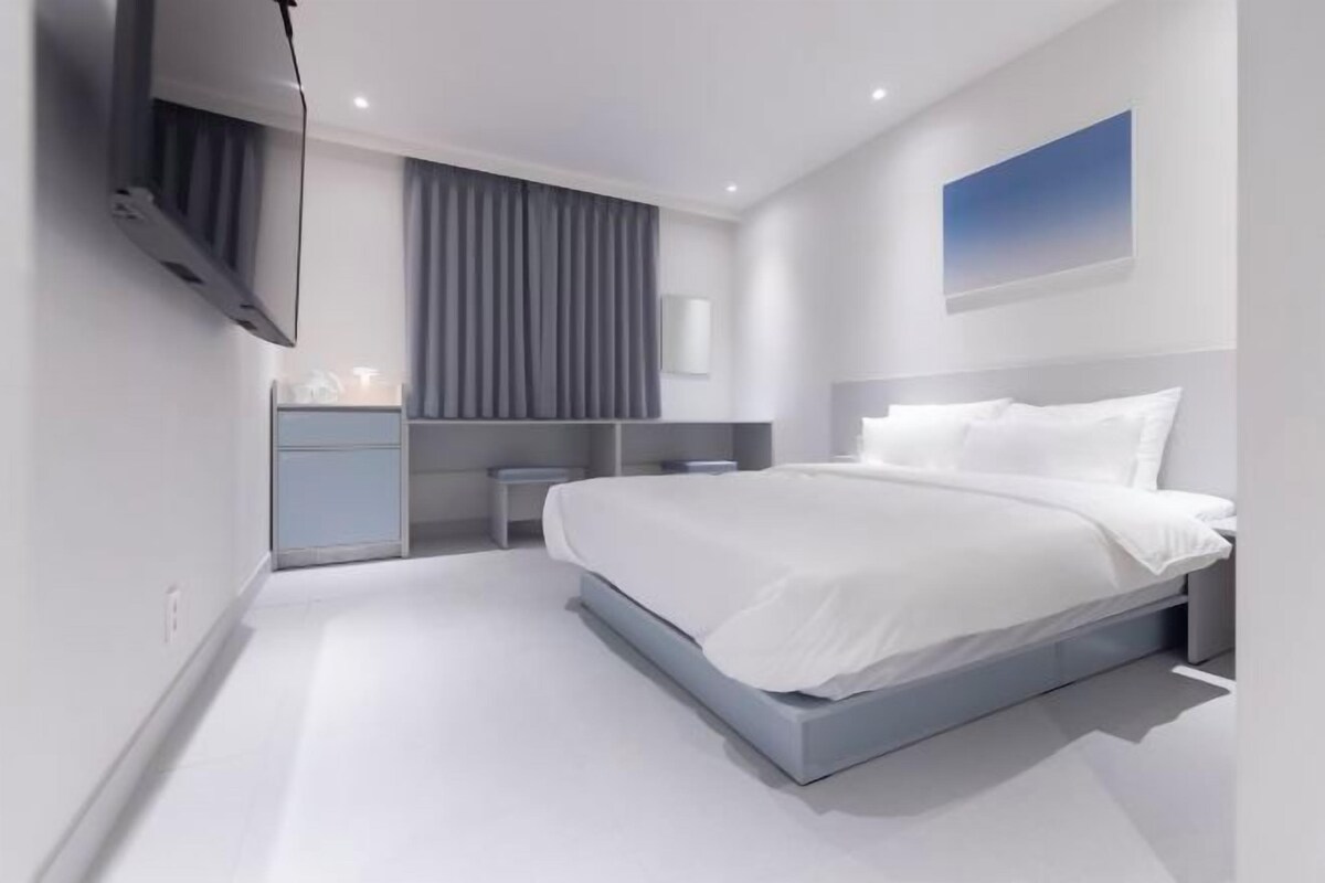 Un dormitorio moderno con una cama grande, una mesita de noche, un televisor montado en la pared y una ventana con cortinas.