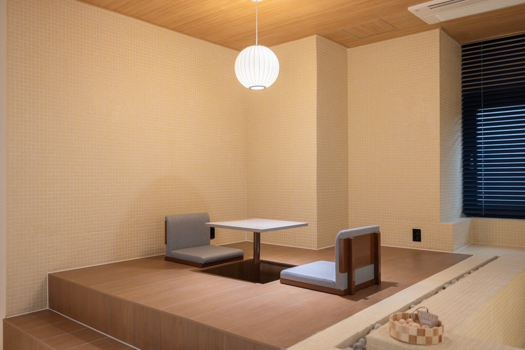 A modern, minimalist room with a wooden floor, a small table, two cushioned seating areas, and a large window with blinds.